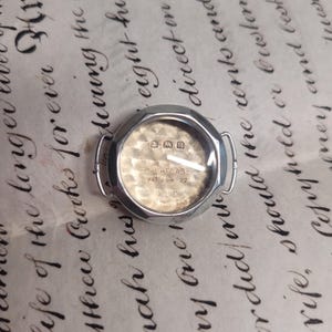 May include: A silver watch with a round face and a silver band. The watch face is white with black numbers and hands. The watch has the text "CACHELASE PAT 269 22" on the face.