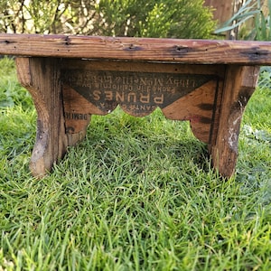 May include: A small, rustic wooden bench with a weathered appearance. The bench has a rectangular seat and four sturdy legs. The front of the bench features a faded label with the word "PRUNES" and other text. The bench is set on a bed of green grass.