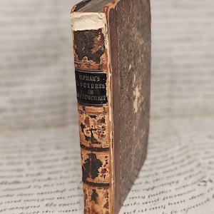 May include: An antique hardcover book with a weathered brown leather spine and mottled brown covers. The spine features a black label with the gold-colored text UPHAM'S LECTURES ON WITCHCRAFT, showing significant wear, frayed edges, and aged paper.