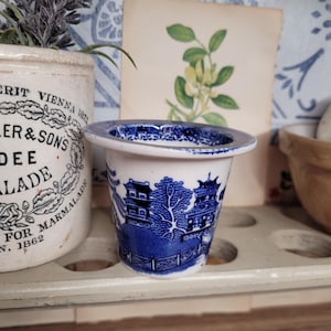 May include: A small, white ceramic cup with a blue and white floral pattern. The cup is decorated with a traditional Chinese design of a pagoda and trees. The cup is sitting on a wooden shelf.