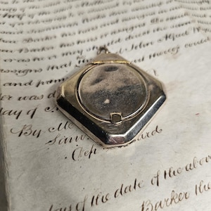 Peut inclure: Un médaillon métallique en forme de losange avec un panneau central circulaire lisse et une petite charnière sur le dessus. Le médaillon présente une surface polie et réfléchissante avec de légers signes d'usure et une finition métallique.