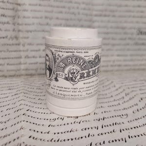 May include: A white ceramic container with a cylindrical shape. The container has a label with the text "LA REINE DES REINES" in black, along with other text and a portrait. The container is likely vintage.