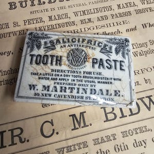 Puede incluir: Antiguo letrero rectangular de pasta de dientes Salicifrice con texto negro sobre fondo blanco. El letrero incluye el texto "Salicifrice, un antiséptico, Tooth Paste" e instrucciones de uso. El letrero está agrietado y envejecido, con una estética vintage.