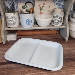 Peut inclure: Un plat de service blanc rectangulaire divisé avec des coins arrondis. Le plat est en céramique ou en porcelaine et repose sur une surface en bois. L'arrière-plan comprend une étagère avec des ustensiles de cuisine et des bocaux vintage.