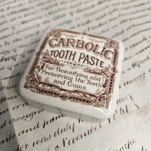 May include: A square, off-white ceramic tile featuring a brown printed label that reads CARBOLIC TOOTH PASTE. For Beautifying and Preserving the Teeth and Gums. The design includes decorative scrollwork borders and a glossy, crackled finish.