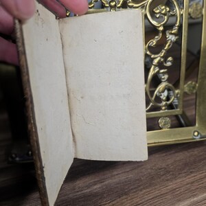 K&ouml;nnte beinhalten: Ein kleines, offenes antikes Buch mit gealterten, cremefarbenen Seiten. Der Einband ist dunkelbraun und abgenutzt. Ein dekorativer Messingrahmen mit kunstvollen Details ist im Hintergrund zu sehen, was auf eine Vintage-&Auml;sthetik hindeutet.