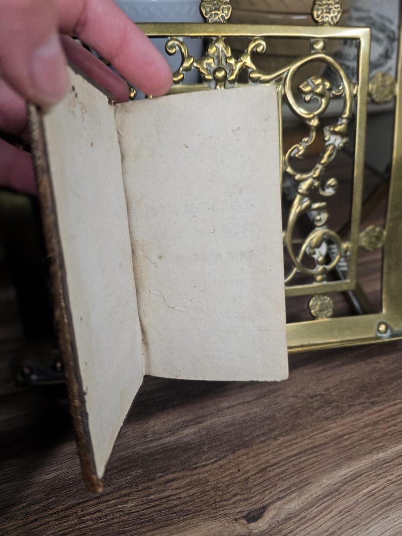 K&ouml;nnte beinhalten: Ein kleines, offenes antikes Buch mit gealterten, cremefarbenen Seiten. Der Einband ist dunkelbraun und abgenutzt. Ein dekorativer Messingrahmen mit kunstvollen Details ist im Hintergrund zu sehen, was auf eine Vintage-&Auml;sthetik hindeutet.