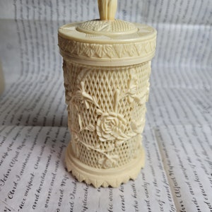 May include: A white, cylindrical, decorative container with a floral design and a lid with a knob. The container is made of a material that resembles ivory.