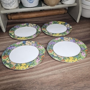 Puede incluir: Cuatro platos ovalados con centro blanco y borde floral con flores amarillas, moradas y verdes. Los platos están colocados sobre una superficie de madera.