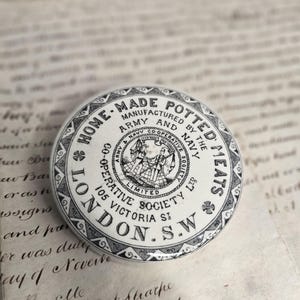 May include: A round white ceramic lid featuring black printed text that reads HOME-MADE POTTED MEATS, ARMY AND NAVY CO-OPERATIVE SOCIETY LTD, 105 VICTORIA ST, LONDON, S.W. The center displays a circular emblem with two figures and a decorative border.