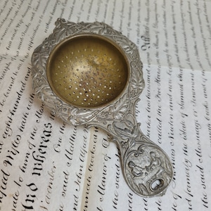 May include: A silver tea strainer with an ornate design featuring floral motifs. The strainer has a round, perforated bowl and a long handle with a detailed depiction of two figures seated at a table.