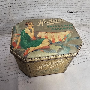 May include: Vintage hexagonal tin box for Swiss Cream Caramels. The lid features an illustration of a woman in a green dress, with the brand name "Heatons" prominently displayed. The box has a gold and green color scheme, with text on the sides.
