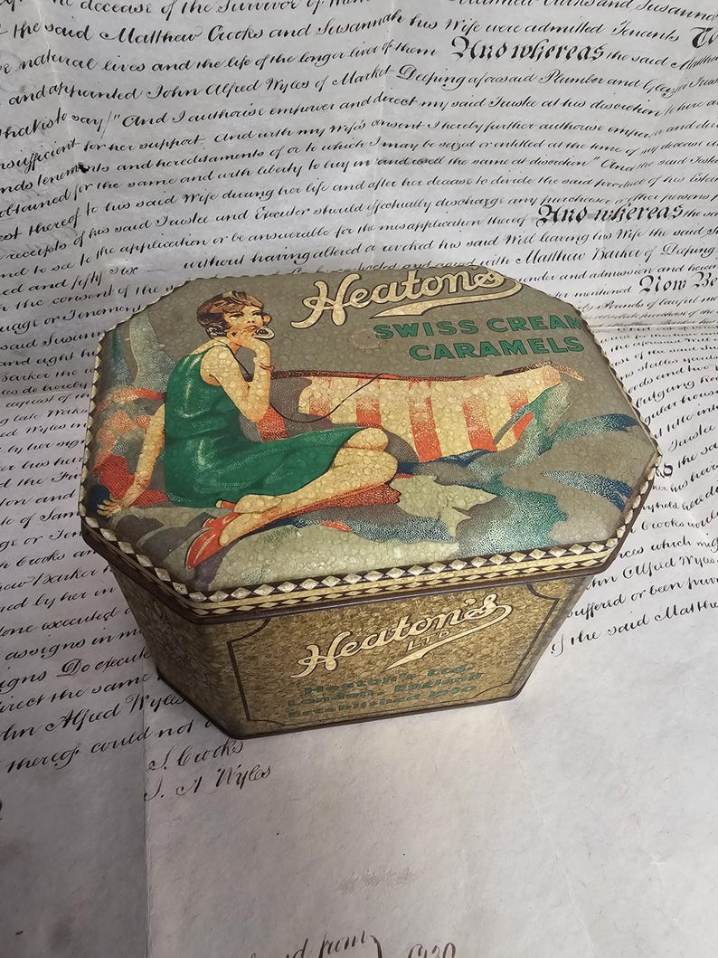 May include: Vintage hexagonal tin box for Swiss Cream Caramels. The lid features an illustration of a woman in a green dress, with the brand name "Heatons" prominently displayed. The box has a gold and green color scheme, with text on the sides.
