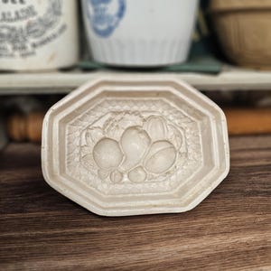 May include: Off-white, octagonal ceramic mold with a fruit design. The mold features a raised relief of pears and other fruits. The background includes vintage ceramic containers and a wooden surface.