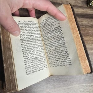 K&ouml;nnte beinhalten: Ein offenes antikes Buch mit gealterten, vergilbten Seiten. Der Text ist auf Franz&ouml;sisch. Das Buch wird von einer Hand offen gehalten, die den gedruckten Text und die gealterten, strukturierten R&auml;nder der Seiten zeigt. Der Einband des Buches ist dunkelbraun.
