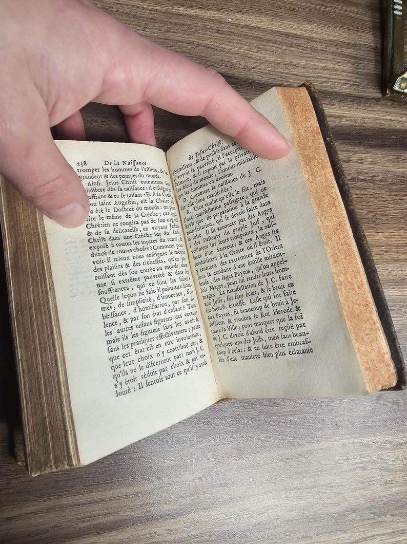 K&ouml;nnte beinhalten: Ein offenes antikes Buch mit gealterten, vergilbten Seiten. Der Text ist auf Franz&ouml;sisch. Das Buch wird von einer Hand offen gehalten, die den gedruckten Text und die gealterten, strukturierten R&auml;nder der Seiten zeigt. Der Einband des Buches ist dunkelbraun.