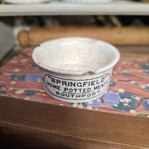 May include: A small, antique white ceramic bowl with a cracked glaze. The bowl has a black printed label that reads "SPRINGFIELD HOME POTTED MEATS SOUTHPORT". The bowl is sitting on a patterned surface.