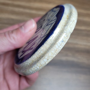 May include: A white ceramic lid with a blue rim and a crackled glaze. The lid has a purple and white design on the inside.