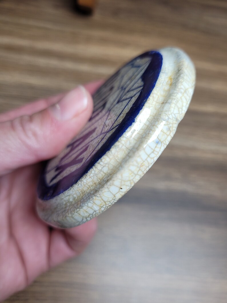 May include: A white ceramic lid with a blue rim and a crackled glaze. The lid has a purple and white design on the inside.