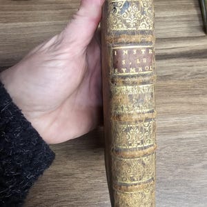 K&ouml;nnte beinhalten: Antikes Buch mit abgenutztem, strukturiertem Einband. Der Buchr&uuml;cken ist hellbraun mit goldenen Filigranarbeiten und dem Titel "INSTR. SYMBOL" in einem rechteckigen Etikett. Das Buch wird von einer Person gehalten.