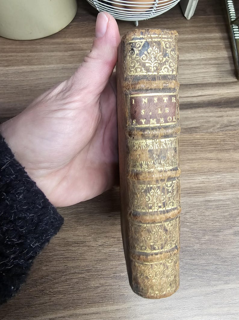 K&ouml;nnte beinhalten: Antikes Buch mit abgenutztem, strukturiertem Einband. Der Buchr&uuml;cken ist hellbraun mit goldenen Filigranarbeiten und dem Titel "INSTR. SYMBOL" in einem rechteckigen Etikett. Das Buch wird von einer Person gehalten.