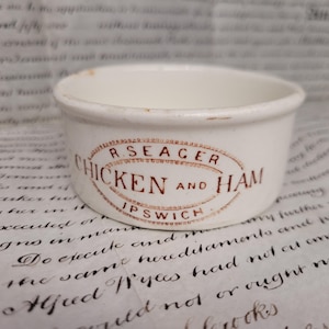 May include: A small, off-white ceramic bowl with a brown oval label that reads "R. SEAGER CHICKEN AND HAM IPSWICH". The bowl has a slightly worn appearance, suggesting age. The background is a vintage paper.