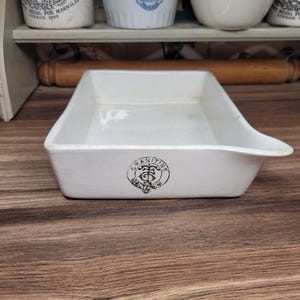 May include: A white rectangular ceramic dish with a pouring spout. The dish has a black emblem that reads "GRANITINE". The dish is on a wooden surface, with other white ceramic containers in the background.