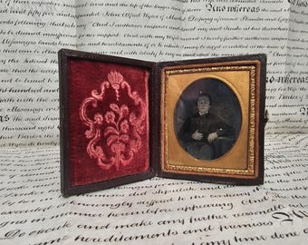 May include: Antique framed portrait featuring a seated person in dark clothing. The portrait is in an oval frame with a gold border, set within a hinged, dark brown case with a red velvet interior.