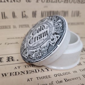 Puede incluir: Un tarro de porcelana blanca con un diseño en blanco y negro. La tapa tiene las palabras "Cold Cream" impresas.
