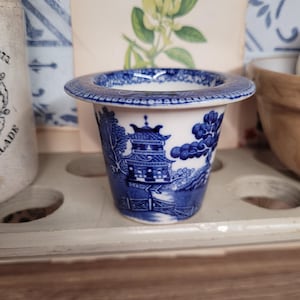 May include: A blue and white ceramic egg cup with a detailed Willow pattern design. The cup features a flared rim and a cylindrical body, showcasing a classic oriental landscape scene. The cup is set against a neutral background.