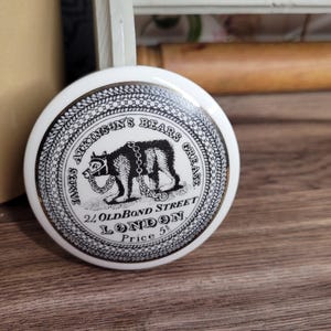 Puede incluir: Una tapa de tarro de cerámica blanca con un diseño en blanco y negro. La tapa presenta un oso con una cadena alrededor del cuello y el texto "James Atkinson's Bears Grease 24 Old Bond Street London Price 5d".