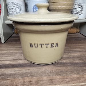 May include: A beige ceramic butter dish with a lid. The word "BUTTER" is printed in dark brown letters on the front. The dish is sitting on a wooden surface. Other ceramic containers are in the background.