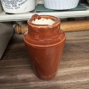 May include: A brown ceramic pitcher with a handle and a spout. The pitcher has a glossy finish and a slightly tapered shape. The word "REGISTERED" is visible on the side. The pitcher is likely vintage and suitable for kitchen decor.