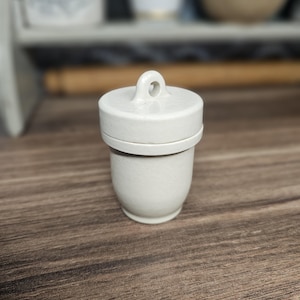 May include: A small, off-white ceramic lidded jar with a smooth, glossy finish. The container features a cylindrical body that tapers slightly towards the base and a matching lid topped with a small, integrated circular loop handle for lifting.