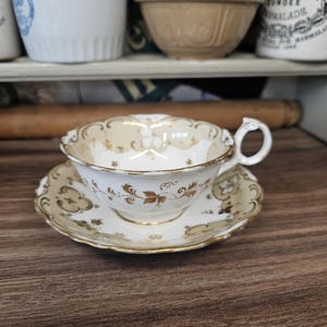 Puede incluir: Un juego de taza y platillo vintage. La taza y el platillo de porcelana blanca presentan diseños florales dorados y ribetes. La taza tiene un asa delicada. El juego se exhibe sobre una superficie de madera oscura.