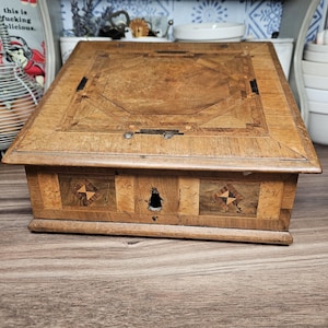 May include: A vintage wooden box with intricate inlaid patterns on the lid and front. The box is rectangular with a keyhole and decorative starburst designs. The wood has a warm, aged tone, suggesting antique craftsmanship.