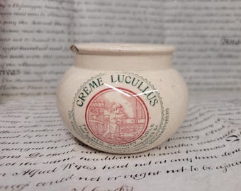 May include: Cream-colored ceramic jar with the text "CREME LUCULLUS" in a red and green circular design. The jar has a rounded shape and a small rim. The label features an illustration of a person cooking.