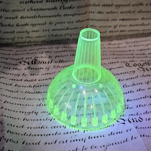 May include: A neon green glass funnel with a flared base and a ribbed, cylindrical neck. The funnel has a series of vertical ridges and a series of small, pointed protrusions around the base. The object is set against a backdrop of aged paper with handwritten text.