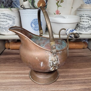 May include: A copper and brass coal scuttle with a lion head design. The scuttle has a handle and a spout. It is sitting on a wooden surface.
