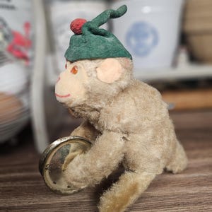 Op de afbeelding: Een vintage knuffelapen-speelgoed met een groene hoed en een rode pompon. De aap houdt een klein, rond, zilverkleurig object vast. Het speelgoed heeft bruin haar en staat op een houten oppervlak. Het gezicht van de aap heeft een rode mond en oranje ogen.