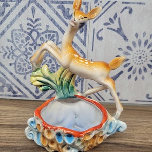 Könnte beinhalten: Eine Keramikfigur eines Hirsches, der auf einem muschelförmigen Pflanztopf steht. Der Hirsch ist braun mit weißen Flecken und hat einen grünen, blättrigen Sockel. Der Topf ist mit blauen, orangen und gelben Details verziert.