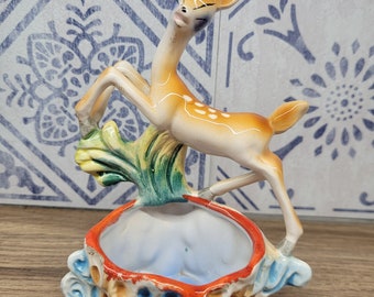 May include: A ceramic figurine of a deer standing on a shell-shaped planter. The deer is brown with white spots and has a green leafy base. The planter is decorated with blue, orange, and yellow details.
