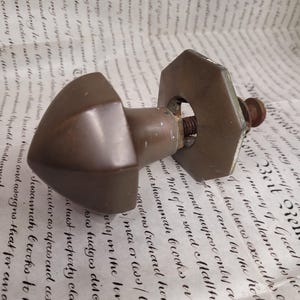 May include: Vintage bronze door handle with a geometric design. The handle has a rounded, triangular shape and is fixed to a hexagonal plate with a threaded bolt. The hardware shows signs of age and wear.