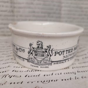 May include: A small, white ceramic bowl with a black and white emblem and text. The bowl has a slightly flared rim and a small handle. The text includes "POTTED MOWBRAY" and "TRADE MARK".