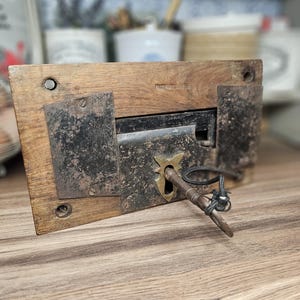 Pode incluir: Uma fechadura de madeira antiga com uma placa frontal de metal e uma chave. A fechadura é retangular com um acabamento escuro, e a chave é inserida na fechadura. A moldura de madeira tem uma cor castanha clara. A chave está presa a um fio preto.