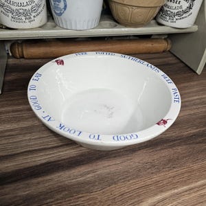 May include: A white ceramic bowl with blue text that reads "GOOD TO LOOK AT. GO TO EAT." The bowl has a wide rim and a shallow interior. The words "PURE POTTED SUTHERLANDS BEEF PASTE" are also visible. The bowl is on a wooden surface.