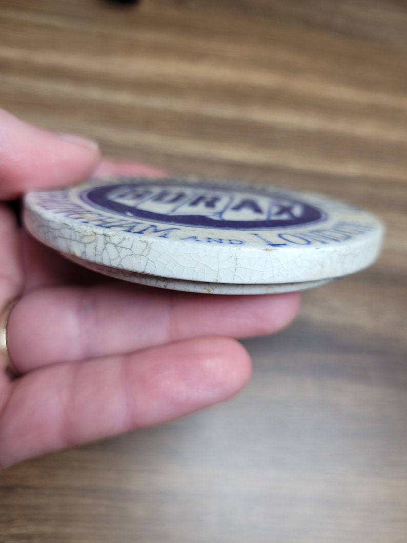 May include: A white ceramic lid with blue lettering that reads "Borax" and "The 20 Mule Team" with a cracked surface.