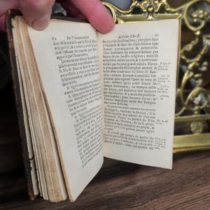 K&ouml;nnte beinhalten: Ein altes Buch mit vergilbten Seiten wird ge&ouml;ffnet und zeigt gedruckten Text. Das Buch hat einen abgenutzten, braunen Ledereinband und befindet sich vor einem dekorativen Messinghintergrund. Die Seiten sind mit detaillierter Schrift gef&uuml;llt.