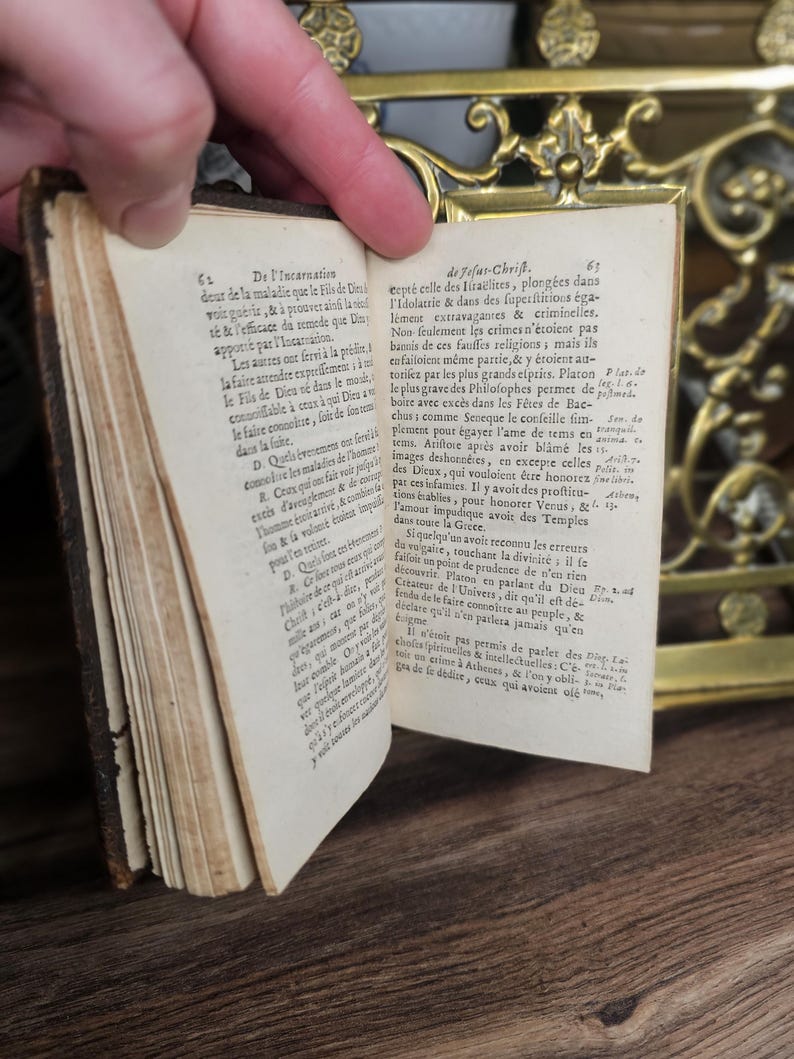 K&ouml;nnte beinhalten: Ein altes Buch mit vergilbten Seiten wird ge&ouml;ffnet und zeigt gedruckten Text. Das Buch hat einen abgenutzten, braunen Ledereinband und befindet sich vor einem dekorativen Messinghintergrund. Die Seiten sind mit detaillierter Schrift gef&uuml;llt.