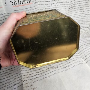 May include: An antique, octagonal tin with a gold interior and a green and gold exterior. The tin is held over a document with handwritten text. The word "Hereas" is visible on the document.